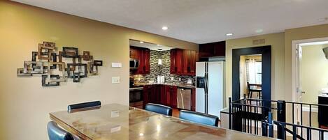 A dining area with a glass-top table and blue chairs, a kitchen with stone backsplash and wooden cabinets, and a living area with a black railing.