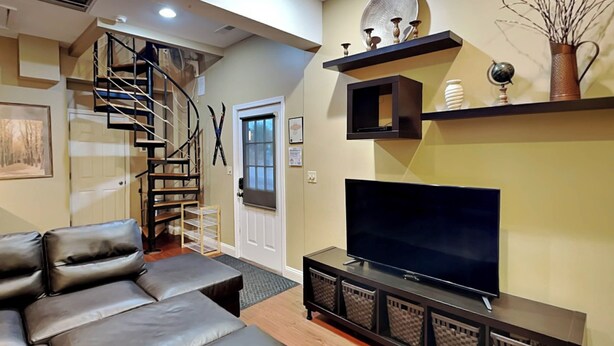 A modern living room with a leather sofa, a flat-screen TV on a wooden console, a staircase, and decorative items on shelves. - Loft of Luxury by Taylor Made Deep Creek (Oakland)