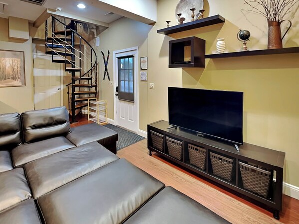 A modern living room with a leather sofa, a flat-screen TV on a wooden console, a staircase, and decorative items on shelves. - Loft of Luxury by Taylor Made Deep Creek (Oakland)