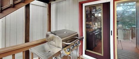 A covered outdoor area with a stainless steel grill, wooden deck, and a glass door leading to an interior space.