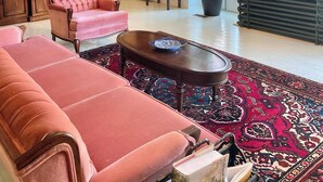 A spacious living room with a large pink sofa, a coffee table, and a chandelier. There is a potted plant and a painting on the wall.