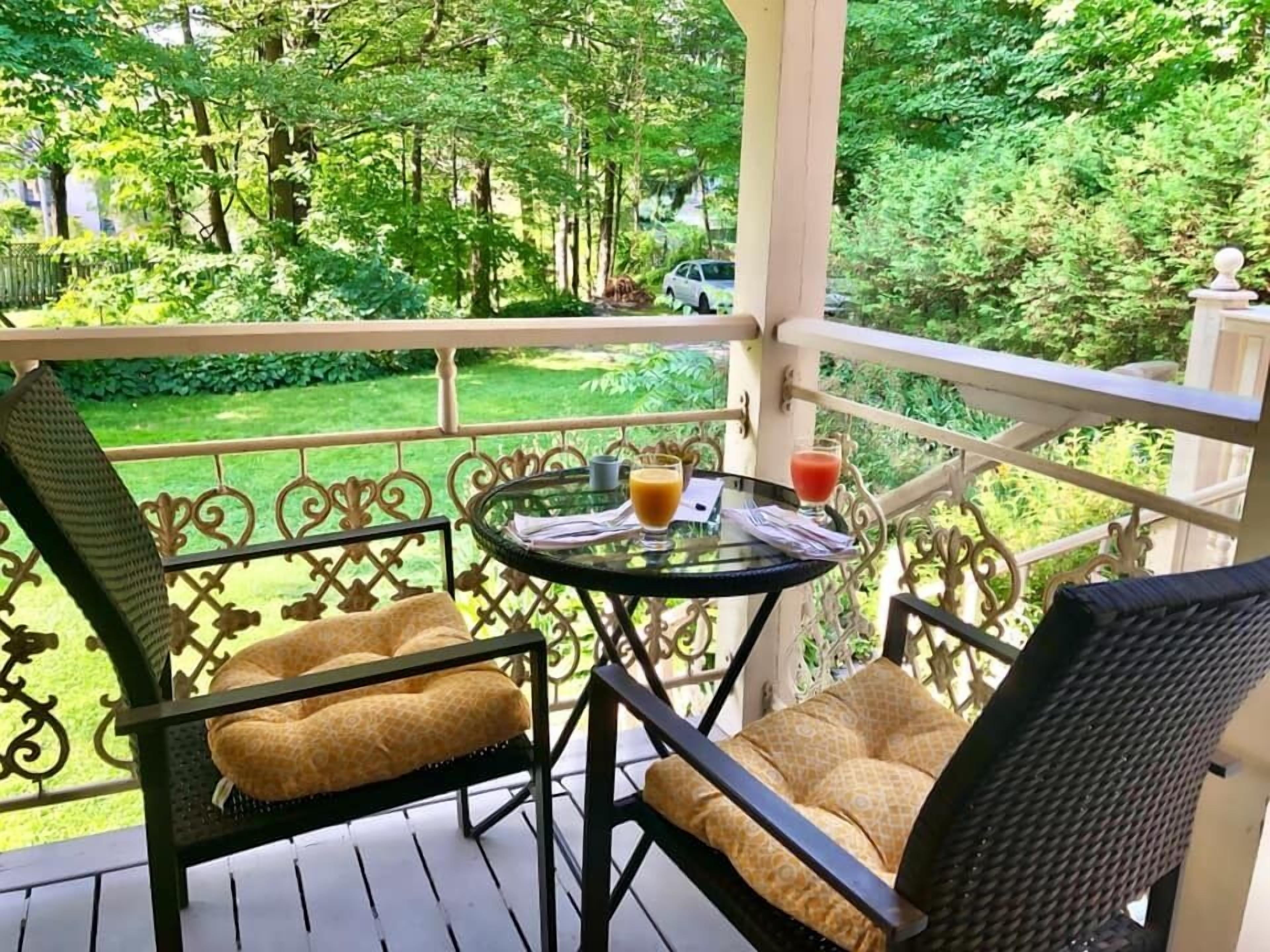 A balcony with wicker chairs and a glass table, two drinks on the table, and a view of a green garden.
