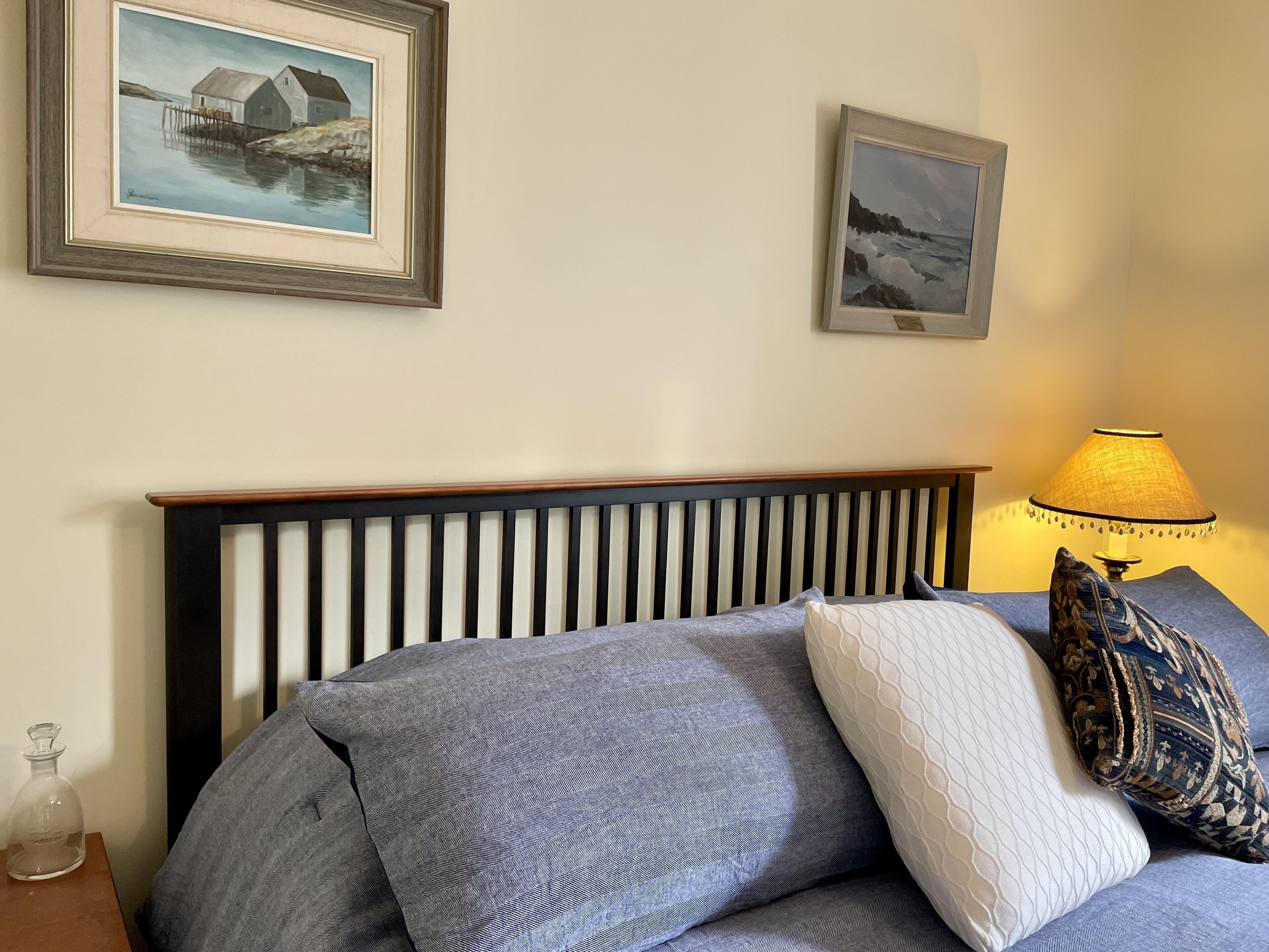 Un letto con testiera in legno, cuscini grigi e bianchi, e una lampada con paralume giallo su un comodino.