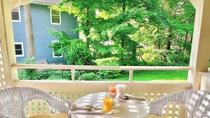 A balcony with wicker chairs, a round table, and a view of a blue house and green trees.
