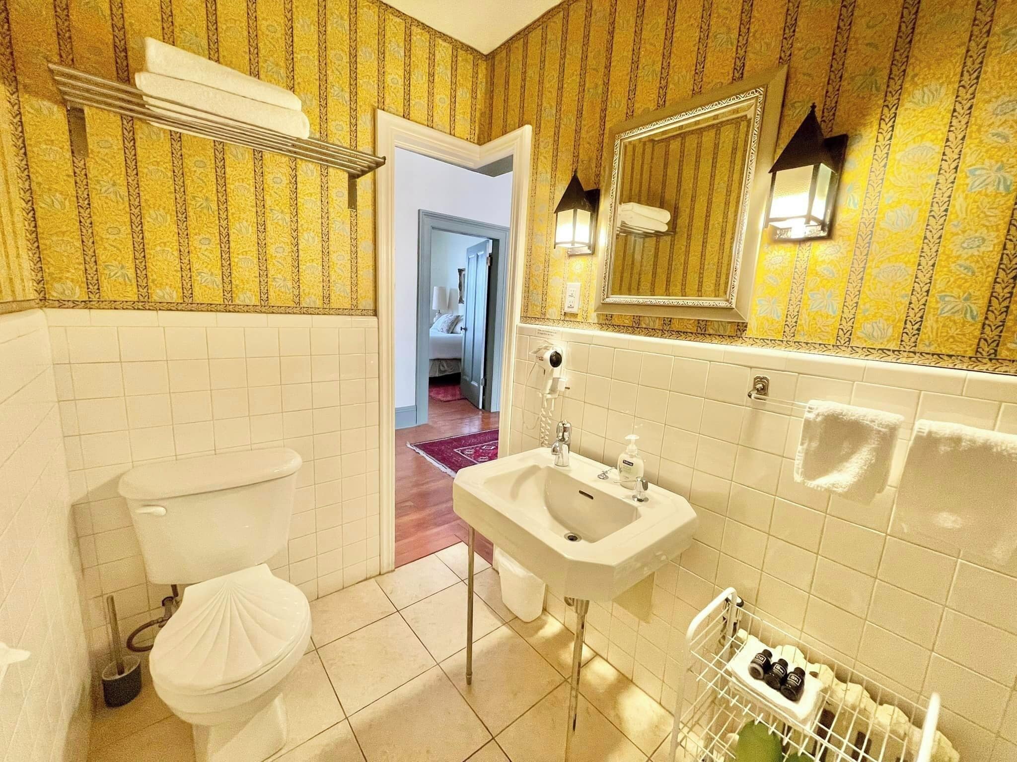 A bathroom with a toilet, sink, and mirror. There is a shower area with a towel rack and a bottle of shampoo. The walls have a patterned wallpaper.