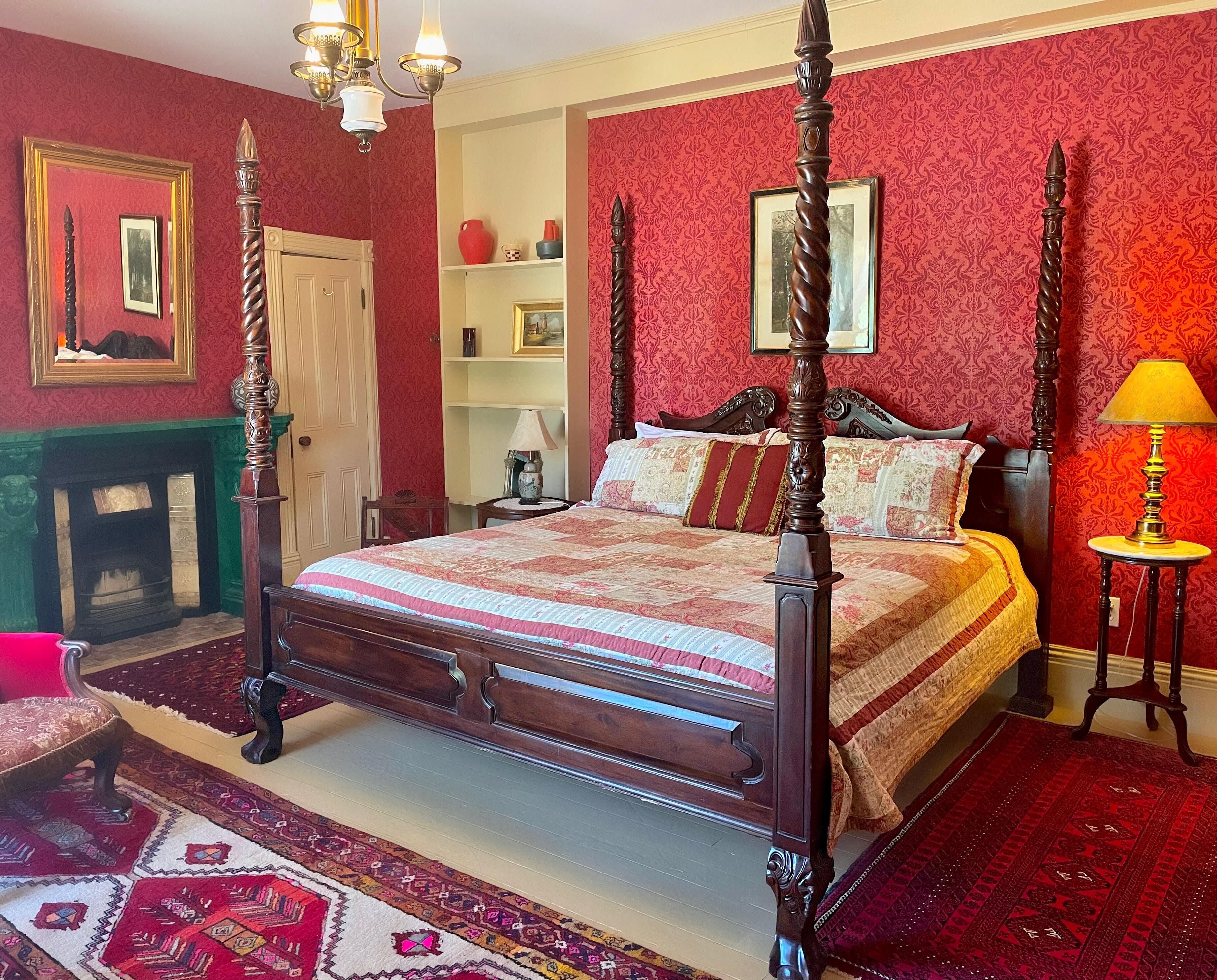 Un lit à baldaquin avec une courtepointe, une cheminée, un mur décoré d’un motif rouge et une petite table avec une lampe.