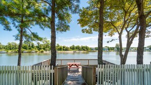 Une terrasse en bois menant à une table de pique-nique située au bord d’un lac, entourée d’arbres et d’une clôture.
