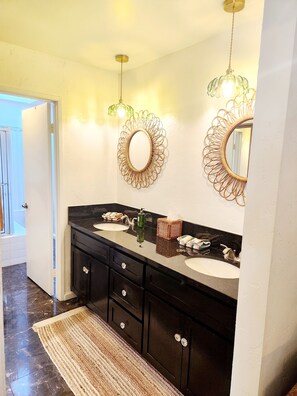 A bathroom with two sinks, dark cabinets, a mirror with a decorative frame, and a green pendant light. - Rancho Mirage/PD border 
28 day min 
3bed, 2bath Pools, tennis/pickleball gated! (Palm Desert)