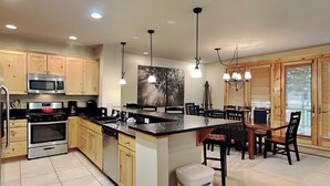 A kitchen with wooden cabinets, a central island, a dining area with a table and chairs, and pendant lighting. - Retreat on Main Street by Taylor Made Deep Creek (McHenry)