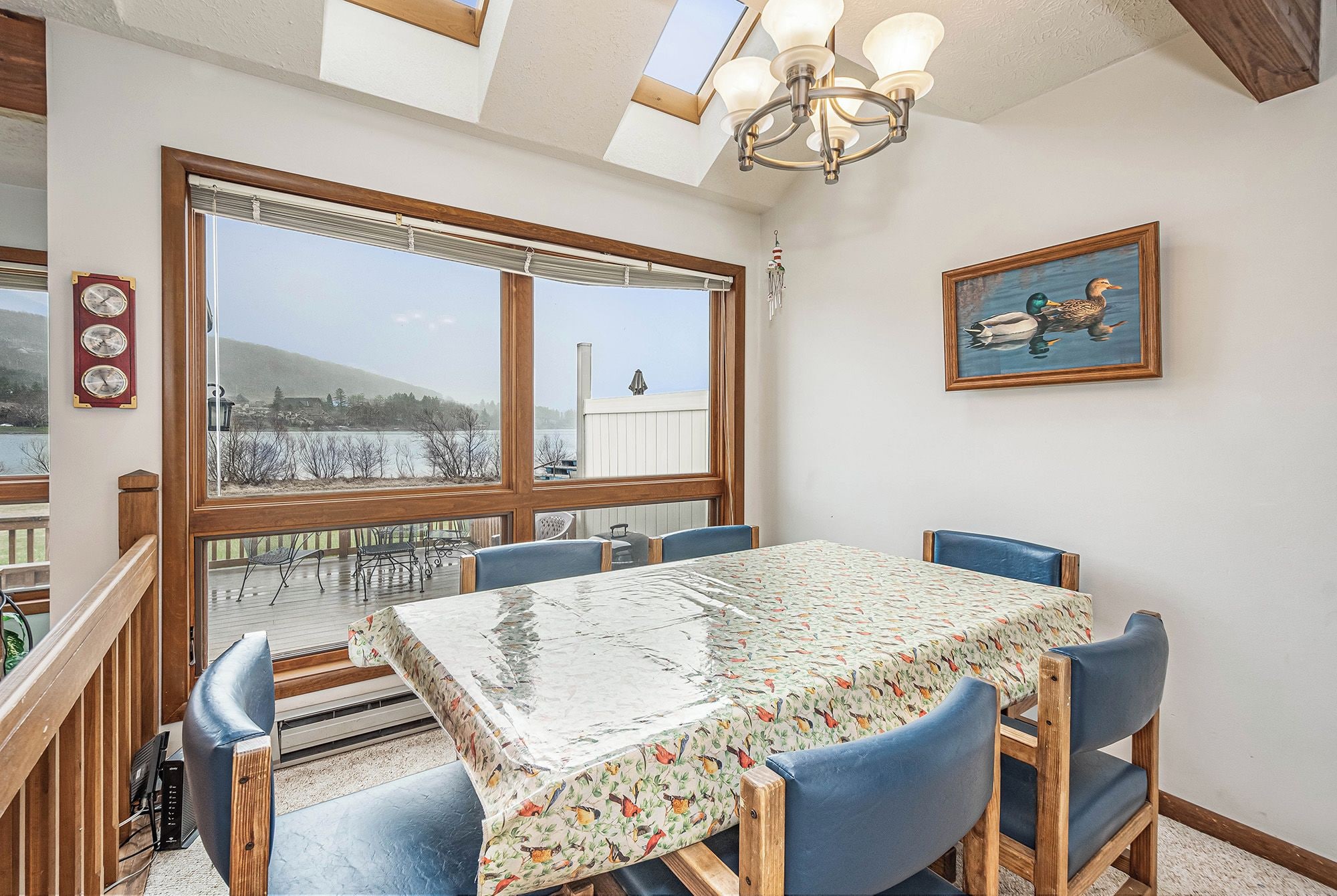 A dining area with a large table, blue chairs, a view of a lake, and a painting of ducks.