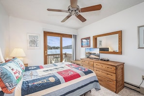 A bedroom with a large bed, a wooden dresser, a ceiling fan, a TV, and a window with a view of a lake.