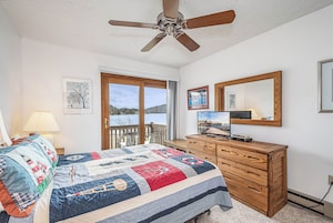 A bedroom with a large bed, a wooden dresser, a ceiling fan, a TV, and a window with a view of a lake.