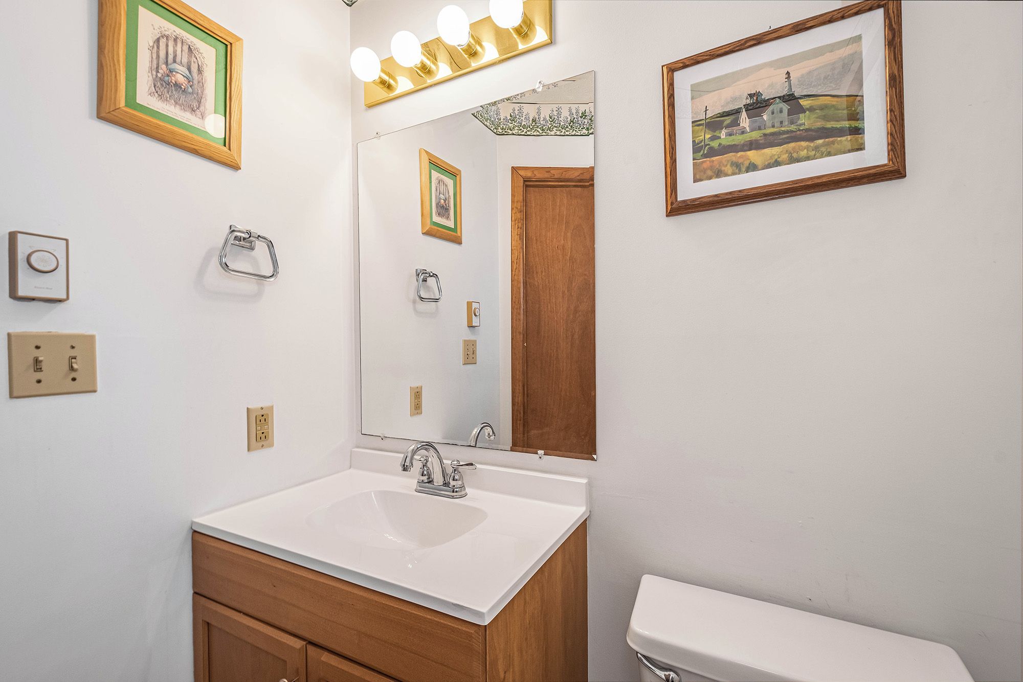 A bathroom with a white sink, wooden vanity, and a large mirror. There are framed pictures on the wall and a light fixture above the mirror.