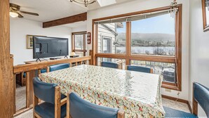 A dining area with a table covered in a floral tablecloth, blue chairs, a ceiling fan, and a view of a house and landscape through large windows. - Ski Harbor 16 by Taylor Made Deep Creek (McHenry)