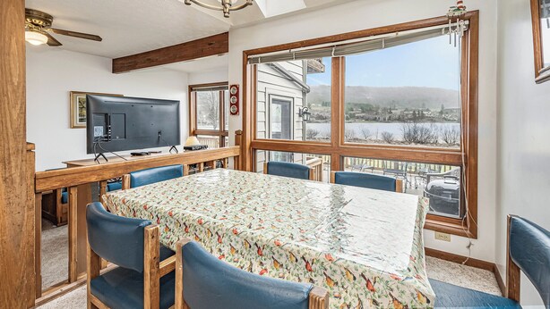 Ein Essbereich mit Tisch, bedeckt mit einer blumigen Tischdecke, blauen Stühlen, einem Deckenventilator und Blick auf ein Haus und die Landschaft durch große Fenster.