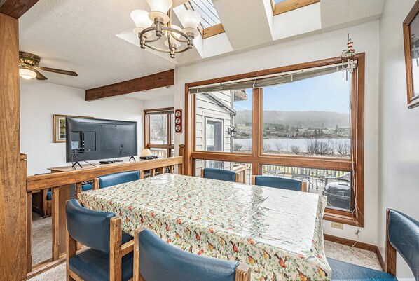 Un coin repas avec une table recouverte d’une nappe à motifs floraux, des chaises bleues, un ventilateur de plafond et une vue sur une maison et un paysage, le tout visible à travers de grandes fenêtres.