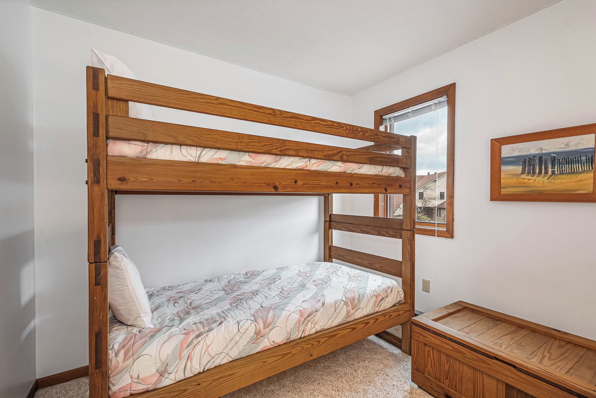 Ein Stockbett mit Holzrahmen und Blumenmuster-Bettwäsche, eine kleine Holzbank und ein Bild an der Wand.