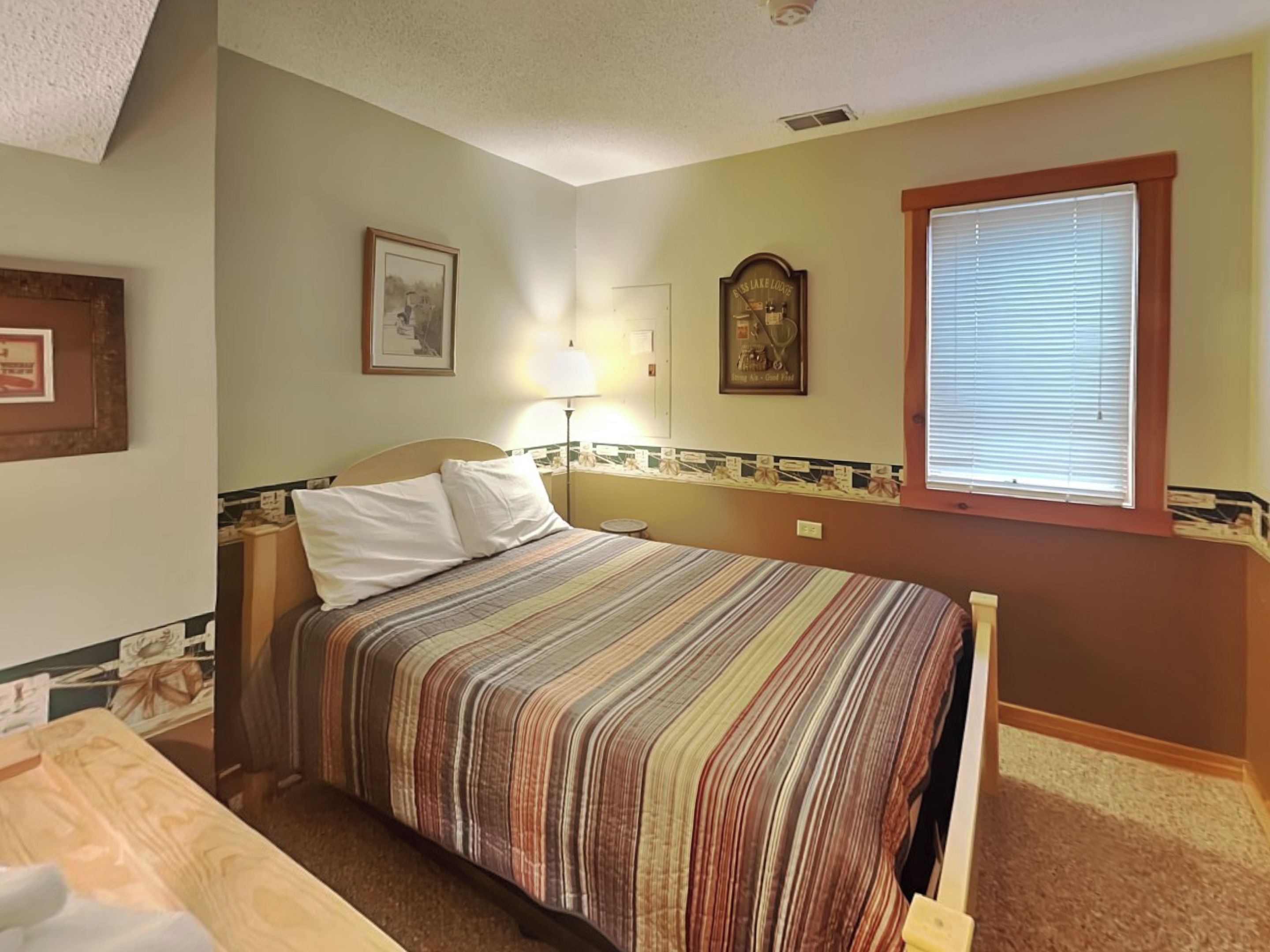 Ein ordentlich eingerichtetes Schlafzimmer mit einem Bett, einem Nachttisch, einer Lampe und einem Fenster mit Jalousien.