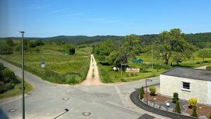 A rural intersection with a dirt path leading to a green field, surrounded by trees and a small building.