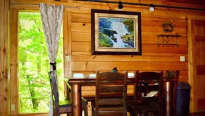 A wooden cabin interior with a dining table, chairs, a framed picture of a waterfall, and a window with a curtain. - ENCHANTED VIEW~**Super cozy & Awesome Valley Views~10 minutes to Pigeon Forge ** (Sevierville)