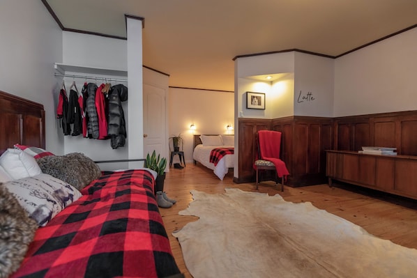 Un dormitorio con una cama, una colcha de cuadros rojos y negros, un manto de piel, un armario de madera y un perchero con chaquetas.