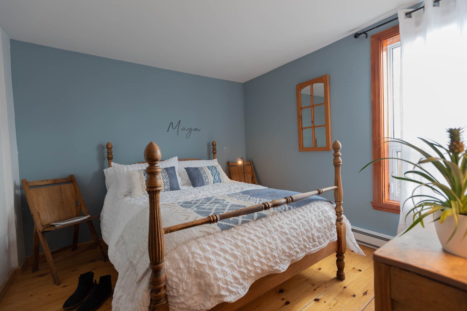 A bedroom with a wooden four-poster bed, a small wooden chair, a bedside table, a potted plant, and a window with a view.