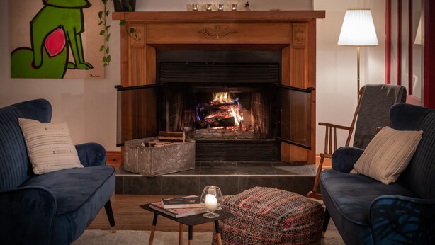 A living room with a fireplace, two blue armchairs, a small table with a candle, and a colorful ottoman. - Auberge et Campagne (Saint Ferreol les Neiges)