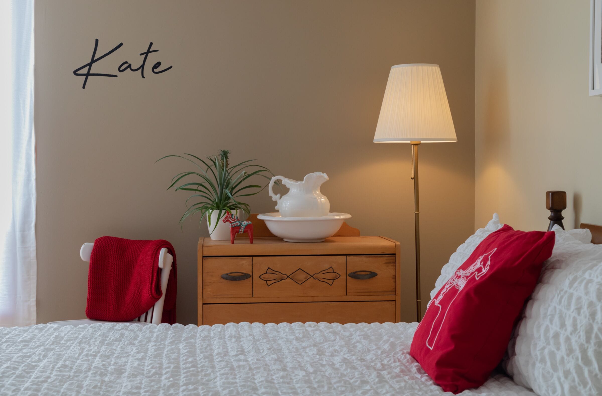 A neatly arranged bedroom with a bed, a bedside table with a lamp, a dresser, and a wall with a name.