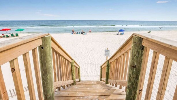 Un escalier en bois mène à une plage de sable où se trouvent des parasols et des chaises de plage.