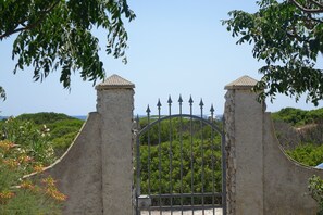 Una entrada porticada con reja metálica y pilares de piedra, rodeada de vegetación y un cielo despejado.