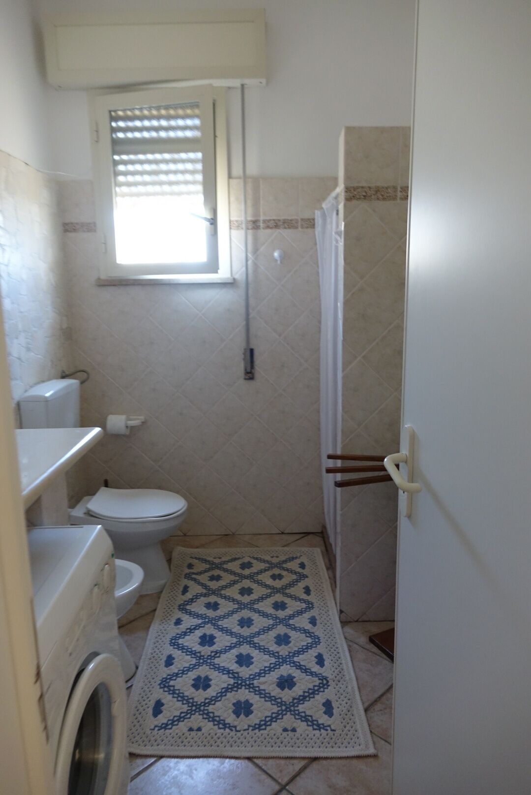 A small bathroom with a toilet, a washing machine, a window, and a shower area.