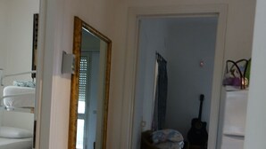 Ein Zimmer mit einem Bett, einem Spiegel, einem kleinen Tisch und einem Stuhl. Eine Tür führt in einen weiteren Raum.