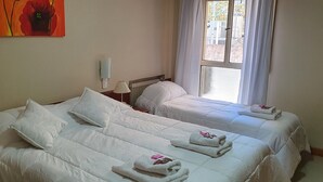 Una habitación de hotel con dos camas, ropa de cama blanca, un cuadro de una flor en la pared, una ventana con cortinas y un radiador.
