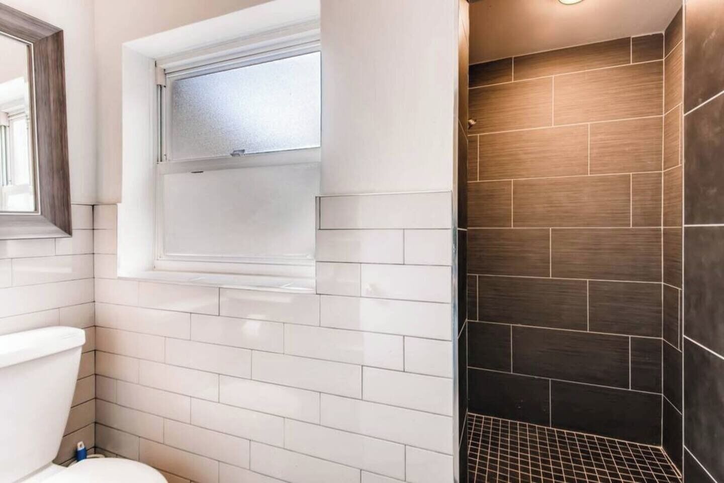 A bathroom with white tiled walls, a window, a toilet, and a shower area with dark tiles.