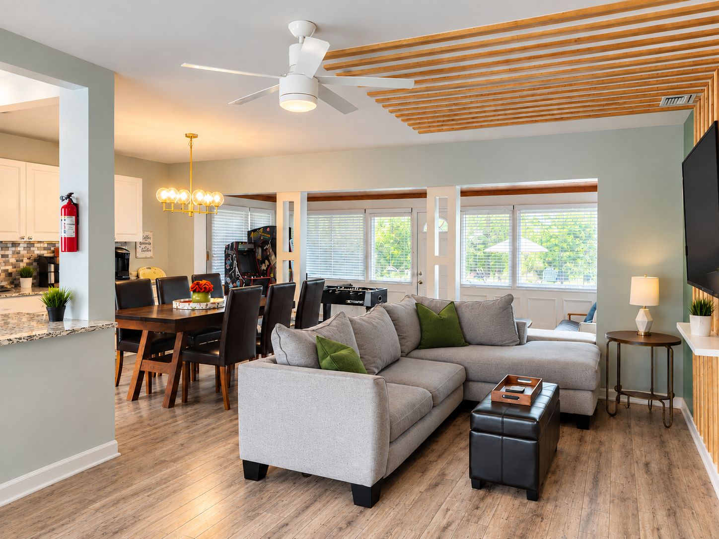 A modern living room with a sectional sofa, a dining area with a table and chairs, a ceiling fan, and a kitchen visible in the background.