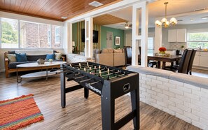 A modern living room with a foosball table, a sofa, and a kitchen area visible through an open doorway. - 7BR Lakefront | Pool & Hot Tub (Orlando)