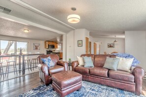 A spacious living room with a brown leather sofa, a matching ottoman, and a blue patterned rug. There is a kitchen in the background with wooden cabinets and a dining area with stools. - Green Acres: Stylish Grill & Chill (Fruita)