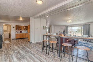 A modern kitchen with wooden cabinets, a central island with stools, and a view of the outdoors through large windows. - Green Acres: Stylish Grill & Chill (Fruita)