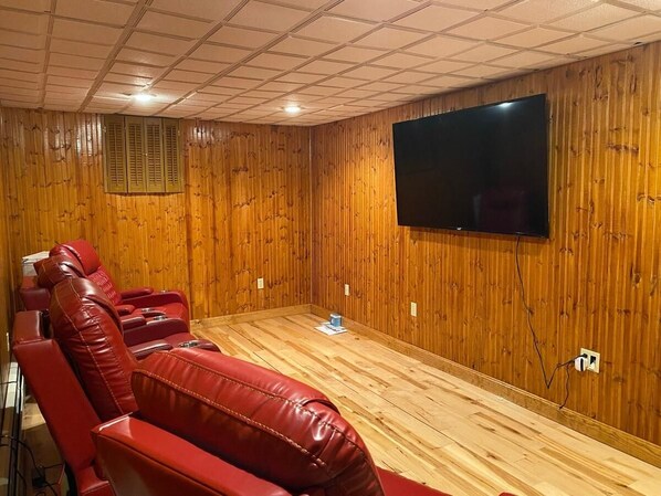 A room with wooden panel walls, a flat-screen TV mounted on the wall, and red leather reclining chairs.