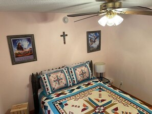 A bedroom with a bed, a ceiling fan, a wall-mounted cross, and framed pictures.
