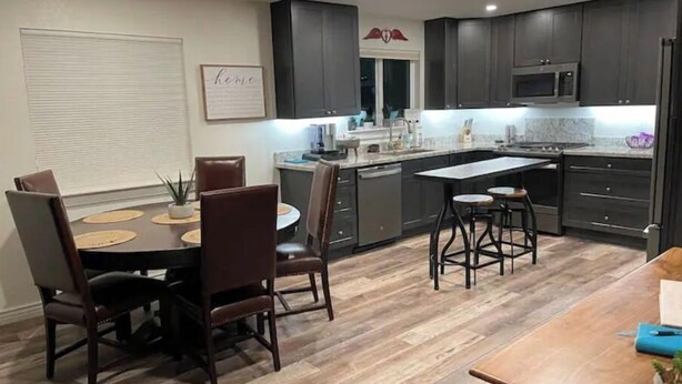 A modern kitchen with dark cabinets, a central island with stools, and a dining area with a table and chairs. - Coffee Pot Rock Views! Equipped with an Electric Vehicle charger. (Sedona)