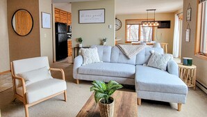 Una sala de estar con un sofá seccional gris, un sillón blanco y una mesa de centro con una planta. Se observa una cocina al fondo con armarios de madera y un refrigerador negro.