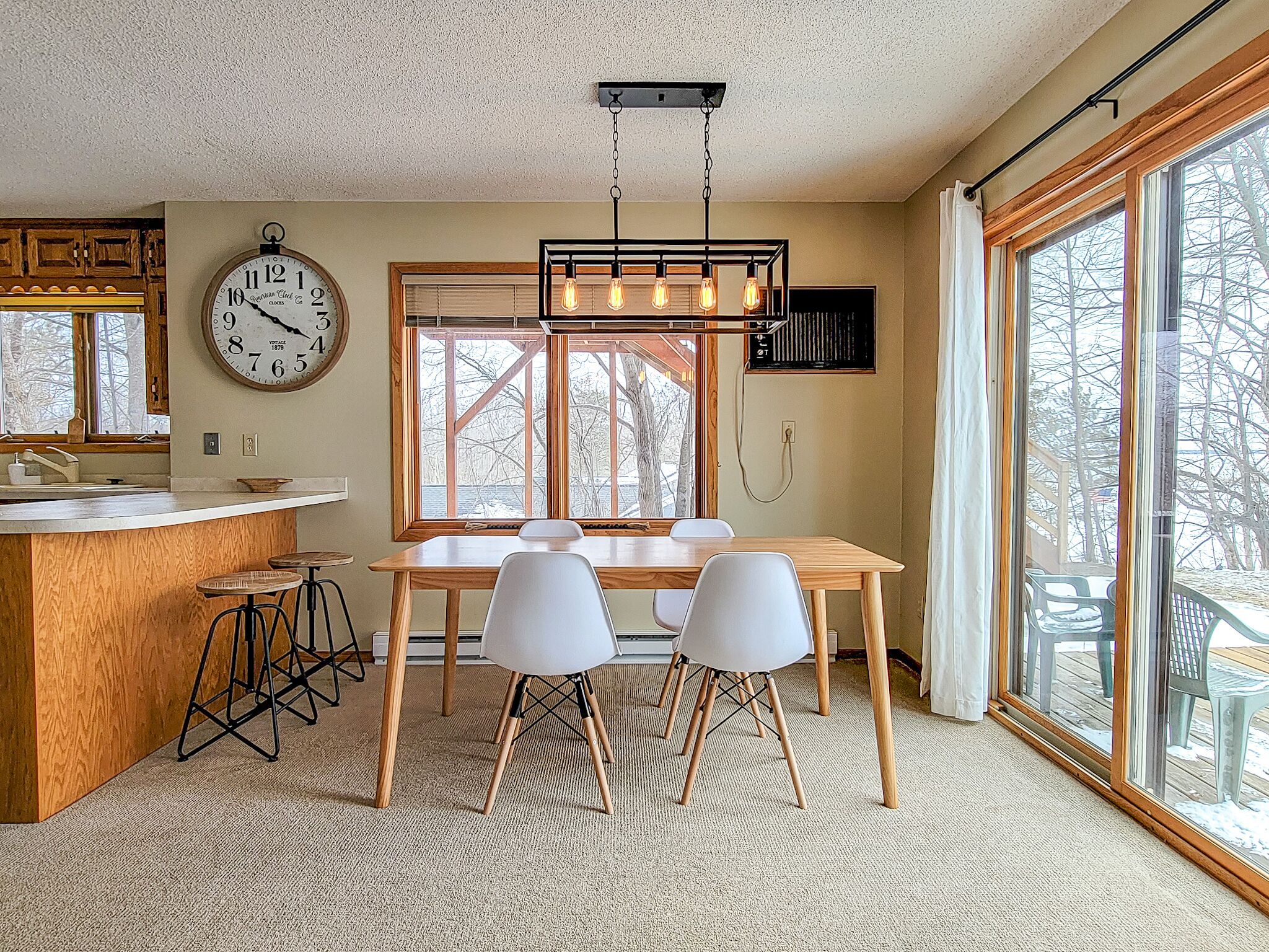 Une cuisine avec un îlot central en bois, une table à manger avec quatre chaises, un luminaire suspendu et une grande fenêtre donnant sur la forêt.