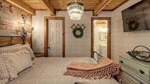 Un'accogliente camera da letto con soffitto in legno, un lampadario, un letto con copriletto decorato, una cassettiera verde e una TV a muro.