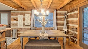 Interno di una baita in legno con un tavolo da pranzo, panche, un lampadario e una grande finestra.