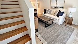 Un escalier en bois mène à un espace de vie comprenant un canapé blanc, une table basse et un tapis.