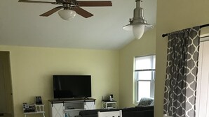 Ein geräumiges Wohnzimmer mit Esstisch, weißen Stühlen, Deckenventilator, Fernseher und einem Fenster mit gemusterten Vorhängen.
