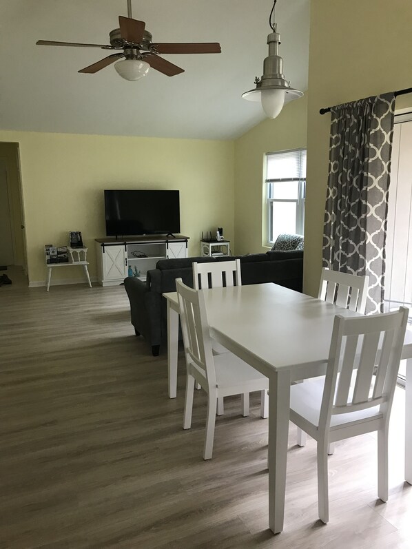 Un salon spacieux comprenant une table à manger, des chaises blanches, un ventilateur de plafond, un téléviseur et une fenêtre agrémentée de rideaux à motifs.