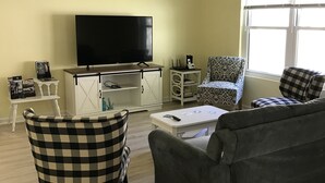 Ein Wohnzimmer mit Flachbildfernseher, grauem Sofa, kariertem Sessel, gemustertem Sessel, einem kleinen weißen Tisch und einem Fenster mit Jalousien.