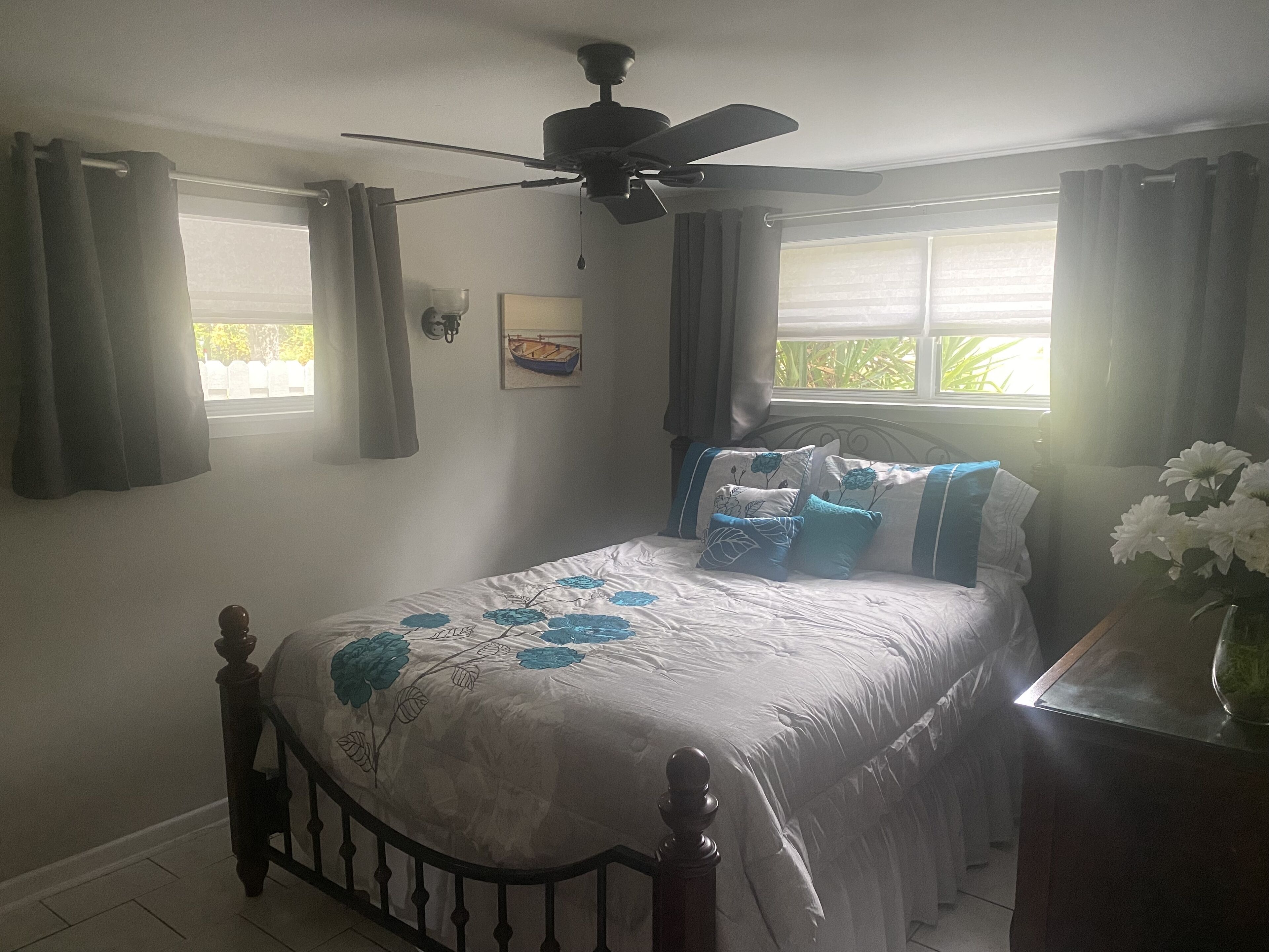 A bedroom with a bed, a ceiling fan, a window with curtains, a wall-mounted picture, and a nightstand with a vase of flowers.